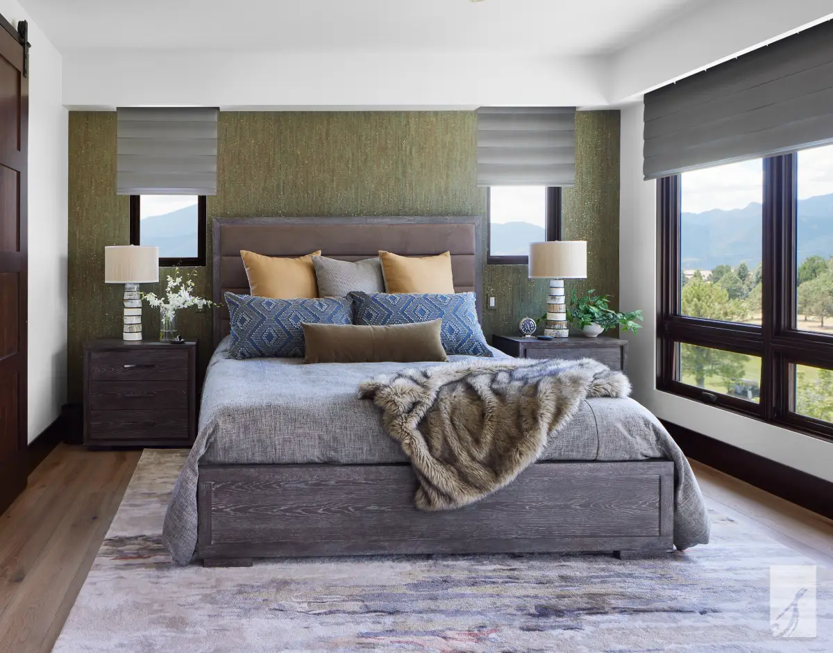 Bedroom with textured green wallcovering, gray Roman shades, and an upholstered channel headboard framed by mountain-view windows