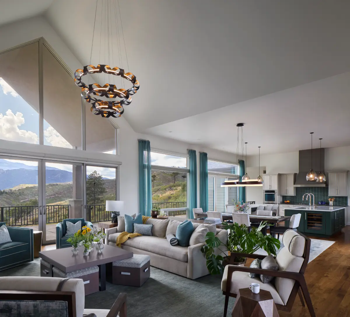 Vaulted great room with mountain views, teal sheer drapery, swivel chairs, and a sculptural tiered chandelier